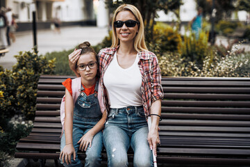 Young blind mother sitting in schoolyard with her little daughter. They smiling and talking...