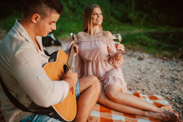 A beautiful caucasian woman in a pastel pink dress  drinking wine and a man playing guitar are sitting on the beach. A loving couple on a picnic by the river