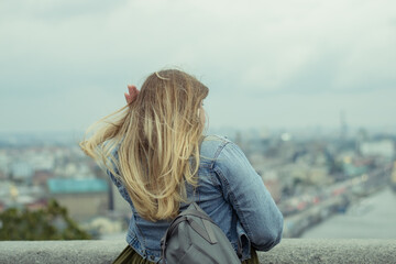 Photo of a young girl with long blond hair from the back in a denim jacket with a backpack on her shoulder. The girl looks at the river.