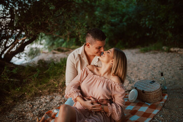 An attractive woman in a pastel pink dress and a man kissing on a blanket on the beach. A loving couple on a picnic by the river