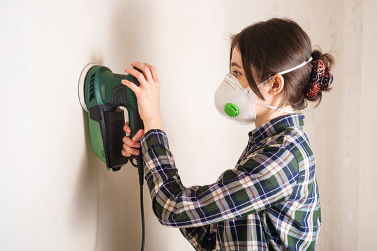 Woman In Protective Mask Working With Electric Sander To Smooth Plaster Wall Surface, Room Renovation Concept