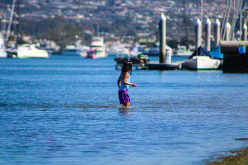 a little girl in a purple bathing suit playing in the ocean water with boats in the harbor at...