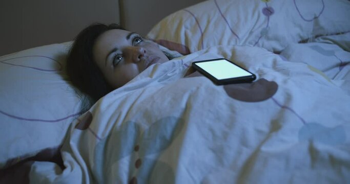 Sad lonely woman ignores calls. A pensive woman, with an apathetic look, lying on the bed, wrapped in a blanket and looking at the ceiling. Night. Phone calling, woman ignoring calls. Seperation