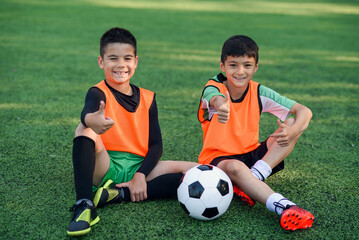 Joyful teenage male soccer players show ok sign resting on artificial turf of football field after...