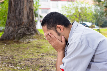 Side shot of young adult man sitting on a city park holding his head with his hands and covering his face.