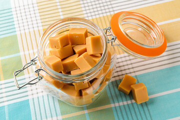 Fresh caramel vanilla fudge candies in an open clip top glass jar over tablecloth. Tasty chewy bonbons made of milk and sugar. Square pieces of delicious soft sweets.