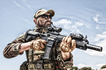 Fototapeta premium Private military company mercenary, brutal looking special forces fighter in battle uniform and plate carrier, wearing radio headset and sunglasses, holding service rifle in hands, ready to fight