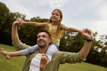 Father Day concept. Little excited girl playing with father in park, sitting on his shoulders and smiling, they are spending time together and having fun