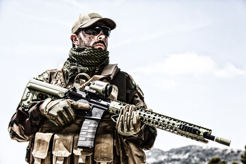 Fototapeta premium Private military company mercenary, brutal looking special forces fighter in battle uniform and plate carrier, wearing radio headset and sunglasses, holding service rifle in hands, ready to fight