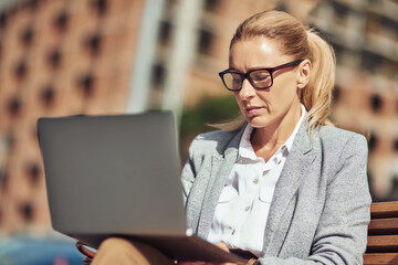 Beautiful middle aged business woman working online, using laptop while sitting on the bench outdoors