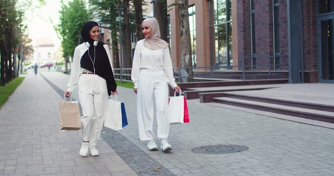 Two confident young Arabic beautiful women in hijabs. Muslim females smiling, discussing, speaking, walking at street of a city, holding shopping bags in hands wearing white headphones on neck.