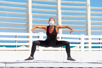 Young woman with protective surgical face mask performing yoga stretching exercises outdoor during...