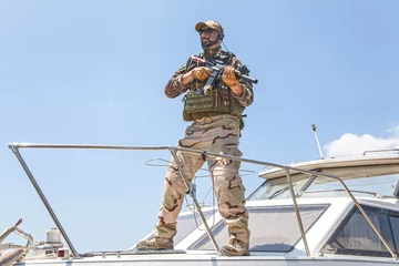 Tuinposter Army special forces soldier, commando fighter in full ammunition, wearing body armor and helmet, armed service rifle, standing on bow of speed boat, looking into distance during seacoast patrolling © Getmilitaryphotos
