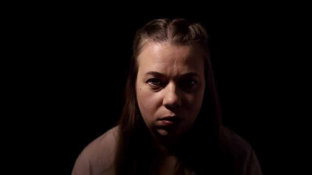 Woman is standing in the studio with black background and speaks aloud. Portrait of caucasian lady with blue eyes talking to someone in darkness. Theme of psychological treatment and psychotherapy.