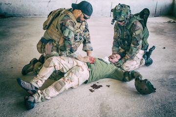 Soldiers trying to stop bleeding at wounded comrade who lying on floor, suffering and screaming in...