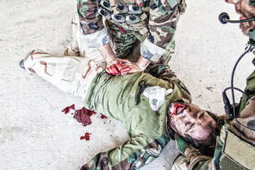 Soldiers trying to stop bleeding at wounded comrade who lying on floor, suffering and screaming in paine. Commando fighter pressing with hands on bloody wound at friends stomach, giving emergency care