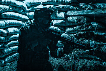 Navy SEALs fighter, army soldier in combat uniform, goggles and battle helmet at night watch, sitting with assault rifle in trench with sandbags, looking in loophole, waiting for enemy attack