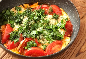 Scrambled eggs with tomatoes, sprinkled with chopped dill and parsley in a teflon skillet on a brown wooden table. Closeup