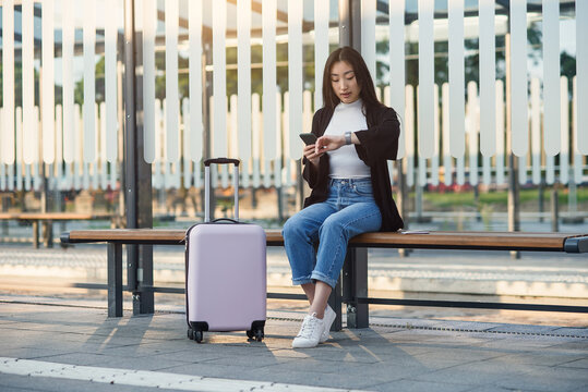 Young asian woman with pink suitcase sits at bus stop and uses cell phone and looks at smart watch.
