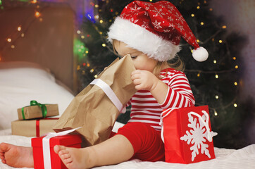 Fototapeta na wymiar Happy Holidays! Adorable baby girl under Christmas tree with gift boxes. Christmas present. Cute girl in Santa hat open gift box.