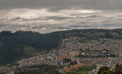 Fototapeta na wymiar Quito, Ecuador - December 2, 2008: Seen from Cerro El Panecillo down. Wide cityscape with towers and regular housing under intense brown and white cloudscape with forested hill on side.