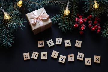 Christmas postcard or background with Christmas tree branches, red berries, gift and wooden letters MERRY CHRISTMAS on dark texture. Festive, holiday concept. Top view, flat lay, selective focus