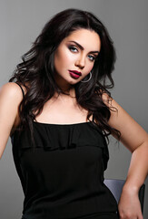 Beautiful makeup woman in black dress with long black curly volume hair style, burgundy lipstick with vamp look on grey background. Closeup sexy