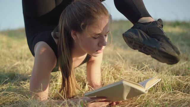 Camera moves down along flexible body of young Caucasian woman in acrobatic pose reading out loud outdoors. Beautiful gymnast combining morning training and hobby at sunrise.
