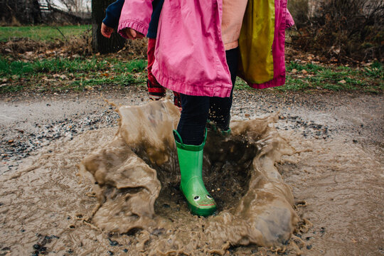 Brother and sister run and splash in puddles in a dirt driveway.