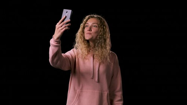 Beautiful nice woman with lollipop in mouth in pink hoodie makes a selfie on a black background. A lady with long blond hair picks up the phone and looks into it, talks to a friend via video link.