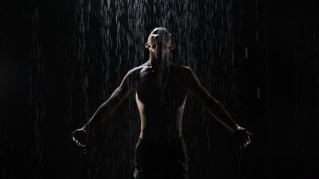 Portrait of pumped-up guy rising hands at night in the rain on black background. Close-up of silhouette of man who is gazing into the camera. Boogie man.
