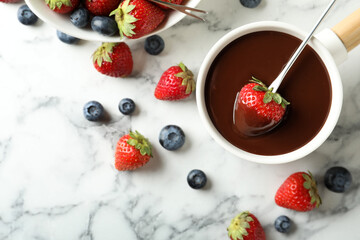 Dipping strawberry into fondue pot with chocolate on white marble table, flat lay
