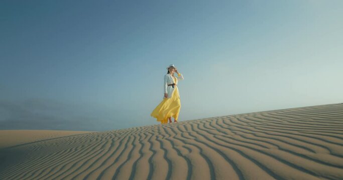 Beautiful fashion footage. Woman in stylish safari outfit is on wavy textured sand desert surface. Slow motion fashion shot with blue sky on background. Advertisement cinematic view of stylish woman