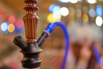 Wooden hookah close-up in the lounge bar on the background of a smoking woman. Abstract bokeh light on background. Traditional Arabian shisha.