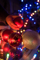 Hand taking a christmas ball over a table with some red and silver christmas balls and christmas lights on the table 