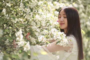 Naklejka premium Beautiful Asian woman in a white dress, standing near a tree with white flowers and holding branches with her hands, looking away