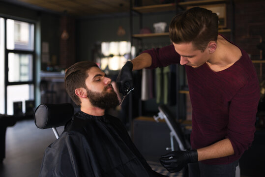 Hairdresser styling customer's beard with scissors at barbershop
