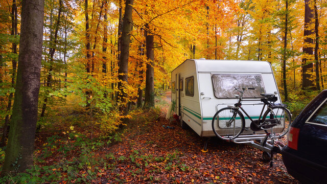 Caravan trailer with a bicycle and a car parked in a golden beech tree forest. Colorful red, orange and yellow leaves on the ground. Autumn landscape. Leisure activity in Heidelberg, Germany