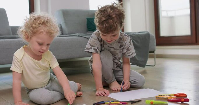 A little boy and a little girl are drawing with pencils on on the white paper on the floor. Looking at each other from time to time. Pencils, paper, kid's scissors and paper knives are in front of