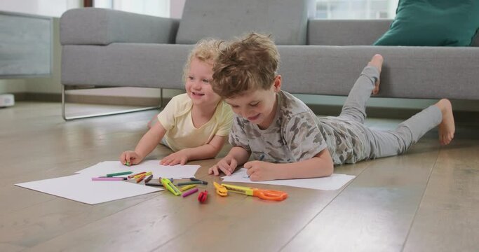 A little boy and a little girl are laying on the floor and drawing with pencils on the white paper, and smiling. Rising heads at their parents and smiling. Pencils, paper, kid's scissors and paper