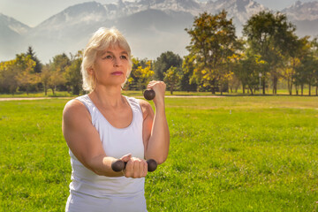 Elderly woman is engaged in fitness with a dumbbell in isolation mode outdoors in a park against a background of mountains on a sunny day. Close-up
