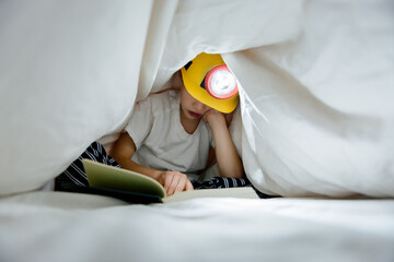 Child hides under bed covers, reading a book by the light of a headlamp
