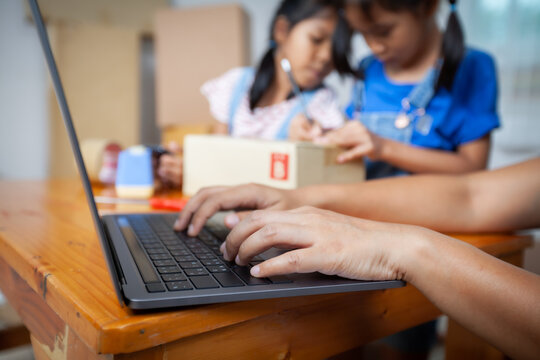 Woman hand using notebook computer taking receive and checking online purchase shopping order. Child is writing address into the box to help parent prepares delivery box for the online customer.