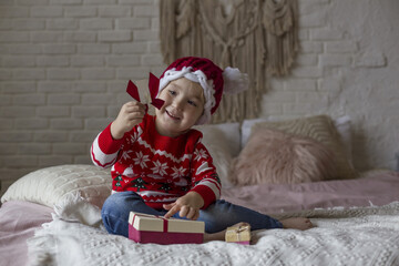 Little girl in a red red christmas sweater and Santa hat plays on the bed with gifts