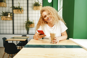 Beautiful curly blonde woman taking selfie while sitting at the table in coffee shop