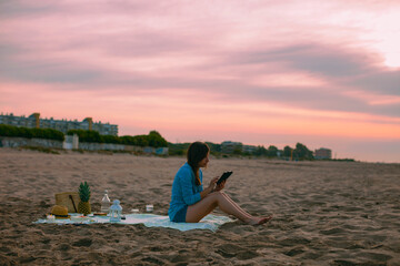 Young beautiful woman reading a book on digital tablet and doing a picnic on the beach at sunset.