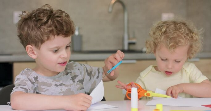 Brother and sister are cutting different objects from paper with scissors, while sitting at the table in the kitchen.