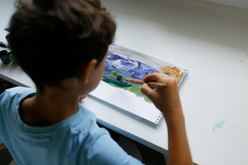 Curly dark-haired boy in a blue T-shirt draws a picture with paints while sitting at a white table