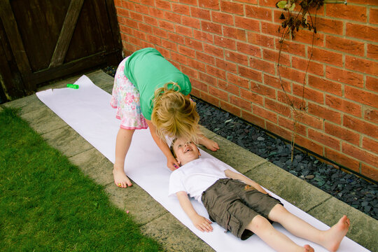 A girl draws the outline of her brother on paper in the garden