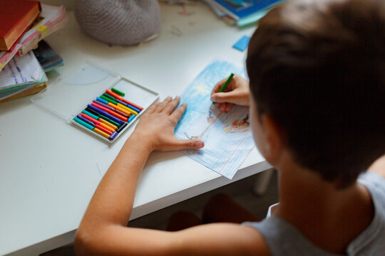dark-haired boy draws a picture with pencils while sitting at a white table